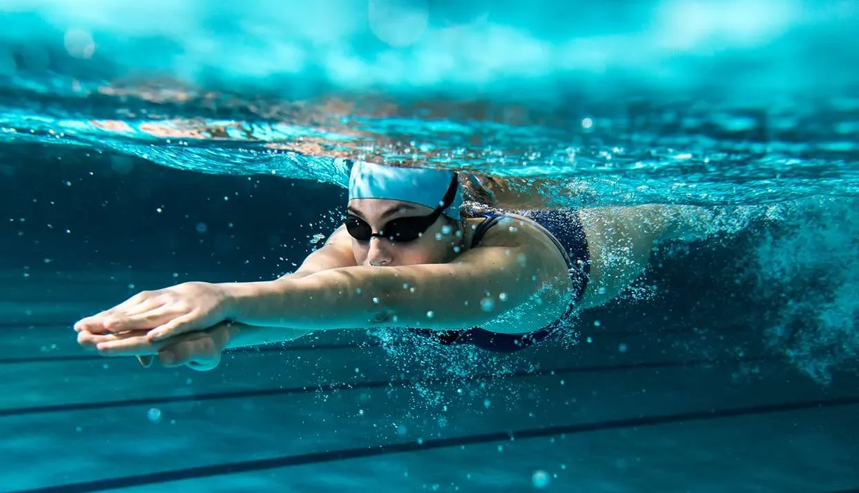 زن ورزشکار با مایو و عینک شنا در حال شنای کرال سینه در استخر به عنوان بهترین ورزش برای لاغری در تابستان